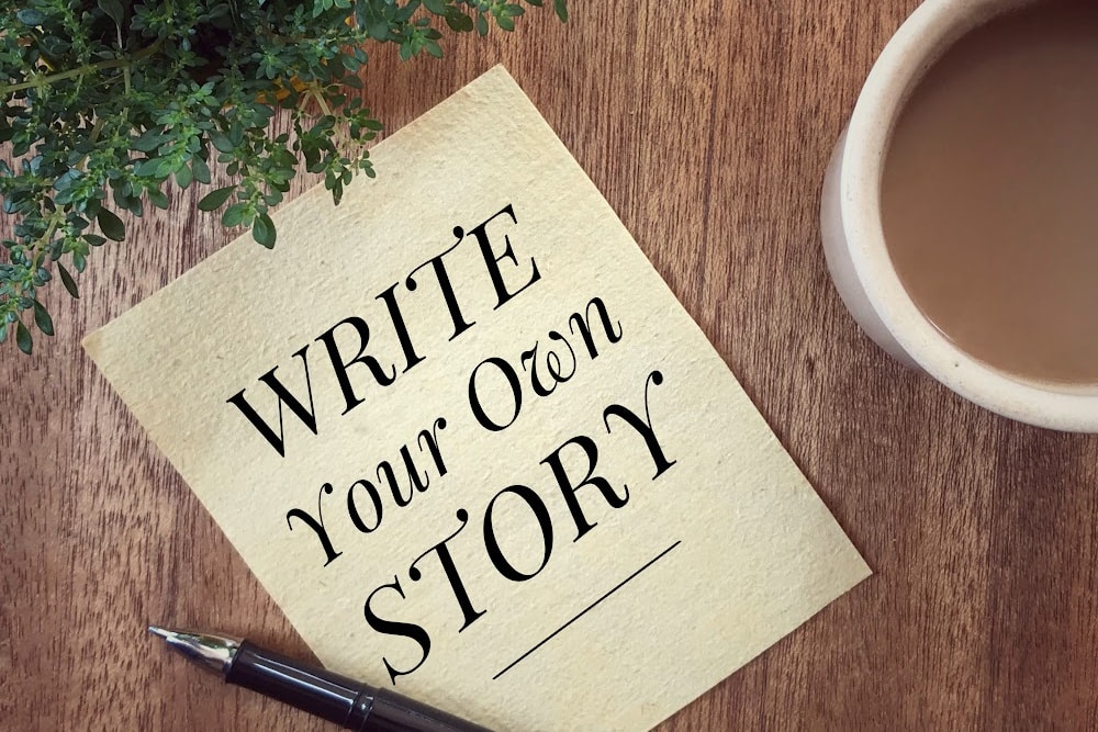coffee and a sheet of paper and pen laying on a table with a plant the words write your own story is on the page.