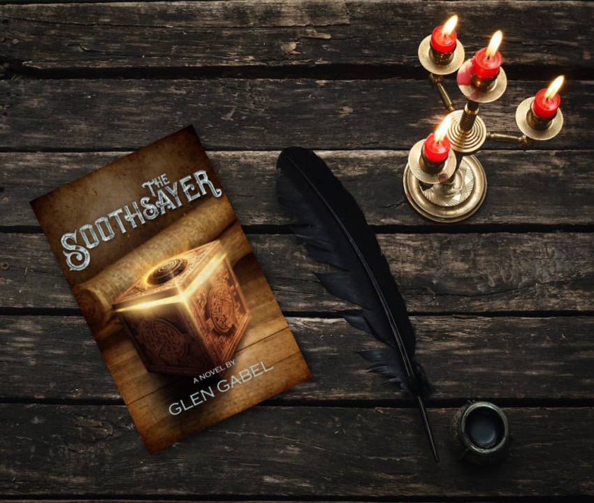 a book The Soothsayer by Glen Gabel is on a table with a quill and a candelabra with red candles that are burning