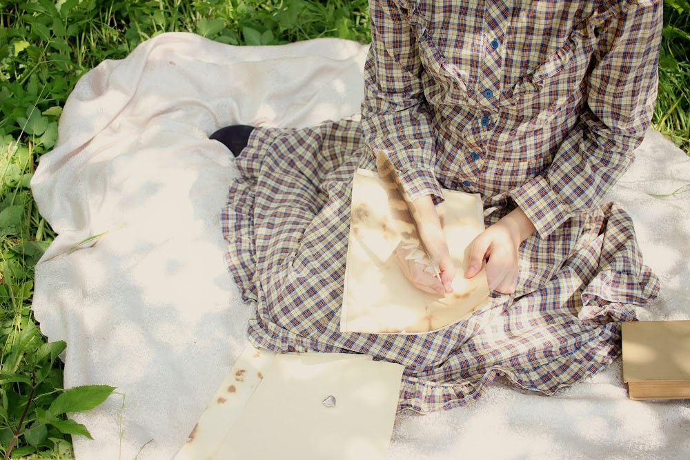 a regency ers woman sitting on a blanket outside writing with a quill