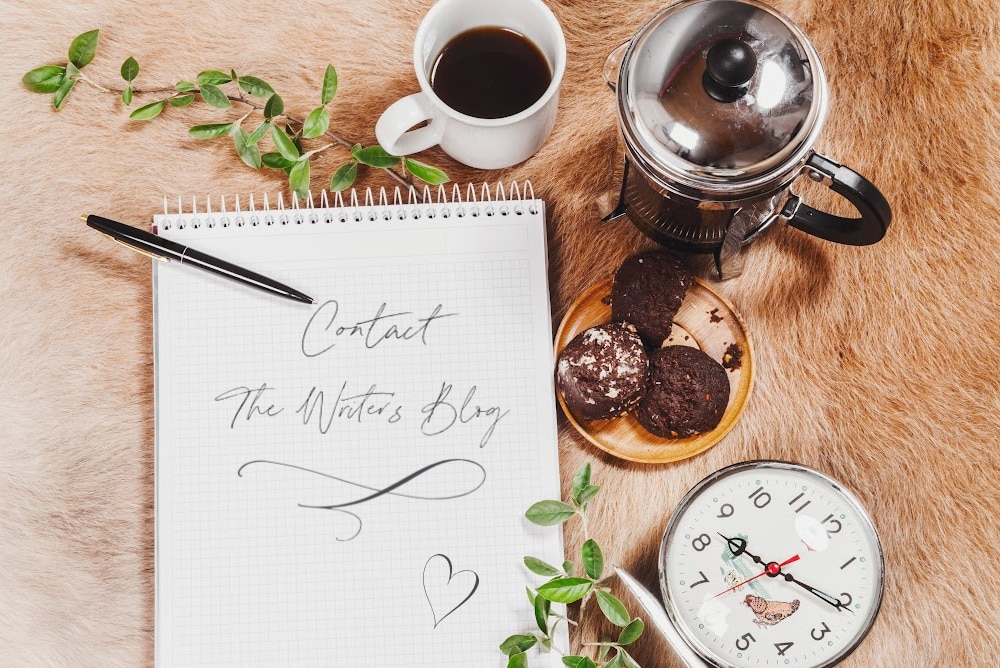 a notebook with contact the writers blog written in calligraphy, a french press coffee cookies and a clock